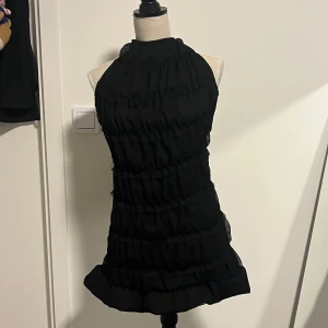 Svart halterneck klänning med volanger - Snygg svart klänning med volanger och knytband i nacken. Perfekt för en elegant kväll. Den har en kort längd och är ärmlös, vilket ger en stilren look.