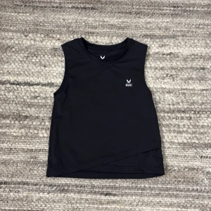 Svart ärmlös topp från SOC - Snygg svart ärmlös topp från SOC med en stilren design. Perfekt för träning eller en aktiv dag. Toppen har en diskret logga på bröstet och en asymmetrisk nederkant som ger en modern touch.😊