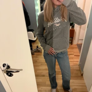 Grå hoodie från Hollister - Säljer en grå hoodie från Hollister med tryck på framsidan. Den har en klassisk känguruficka och en bekväm huva. Perfekt för en avslappnad stil.