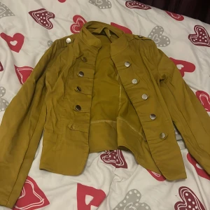 Gul jacka från Tally Weijl - Säljer en snygg gul jacka från Tally Weijl med stora silverfärgade knappar. Jackan har en unik design med en hög krage och långa ärmar. Perfekt för att ge din outfit en färgklick!