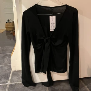 Svart långärmad knytblus från Gina Tricot - Snygg svart långärmad blus från Gina Tricot med knytning framtill. Perfekt för en stilren look. Blusen har en v-ringad design och är tillverkad i ett mjukt material.