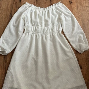 Vit klänning från H&M  - Säljer en söt vit klänning från H&M med elastisk offshoulder-design och långa ärmar. Klänningen har ett diskret prickigt mönster och smockdetaljer i midjan för en fin passform. Perfekt för sommarens alla tillfällen! 🌼