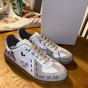 Vit sneaker med handskrivna detaljer från Maison Margiela - Unika vita sneakers från Maison Margiela med handskrivna detaljer i blått och svart. Skorna har en klassisk design med snörning och en kontrasterande brun sula. Perfekt för den som vill ha något annorlunda och stilrent.