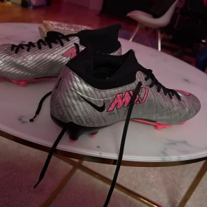 Nike fotbollsskor i silver och rosa - Snygga Nike fotbollsskor i silver med rosa detaljer och svart snörning. Skorna har en modern design med Nike-loggan tydligt synlig på sidan. Perfekta för fotbollsplanen med sina greppvänliga dobbar.