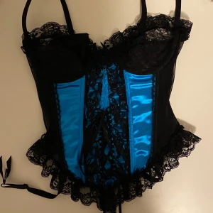 Svart och blå korsettop med spets - Snygg korsett i svart och blått med spetsdetaljer och snörning framtill. Den har justerbara axelband och en volangkant nedtill. Passar strl. XS-S.