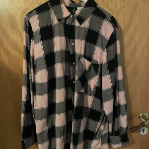 Rutig skjorta från H&M Divided - Snygg rutig skjorta från H&M Divided i svart och rosa. Skjortan har långa ärmar och en klassisk krage. Perfekt för en avslappnad stil med knappar framtill och en bröstficka.