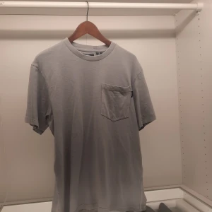 Grå t-shirt från Superdry - Säljer en stilren grå t-shirt från Superdry med en praktisk bröstficka. T-shirten är kortärmad och tillverkad i mjukt material för en bekväm passform. Perfekt för en avslappnad look.