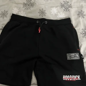 Svarta shorts från Hoodrich - Snygga svarta shorts från Hoodrich med vit och röd logga på benet. De har snörning i midjan för justerbar passform och en liten etikett på sidan. Perfekta för en avslappnad stil. Pris kan diskuteras!!
