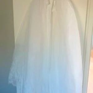 Vit klänning med spets och tiara - Elegant vit klänning med spetsdetaljer och ett vackert bälte med broderad dekoration. Klänningen har en lång, flödande kjol och är perfekt för speciella tillfällen. Medföljer en glittrig tiara som ger en extra touch av glamour.