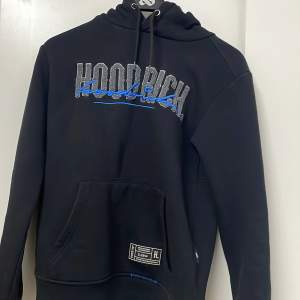 Säljer en svart hoodie från Hoodrich i storlek XS. Den har en cool design med texten 'Hoodrich' i grått och blått på framsidan. Hoodien har en stor ficka framtill och en justerbar huva. Perfekt för en avslappnad stil!