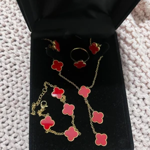 Smyckesset  - Smyckesset med armband, halsband, ring och örhängen. Setet är designat med röda klöverformade hängen på en guldfärgad kedja, vilket ger en lyxig och stilren känsla. Perfekt för att komplettera en festlig outfit eller för att ge bort som en present. S