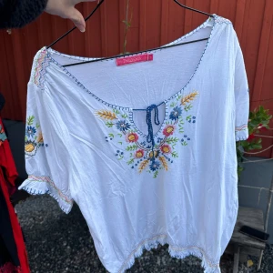 Vit blus med broderade blommor - Säljer en superfin vit blus från Indiska med färgglada broderade blommor på framsidan och ärmarna. Blusen har korta ärmar och en söt knytning vid halsen. Perfekt för en somrig look!