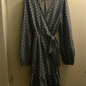 Klänning - Säljer en superfin blå klänning med vita prickar. Den har en omlott-design med knytband i midjan och volangdetaljer längst ner. Den är långärmad , perfekt för både vardag och fest. Materialet känns lätt och bekvämt. ( justerbar, funkar för s eller L)
