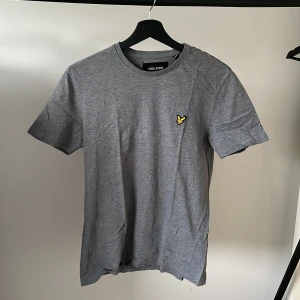 Grå t-shirt från Lyle & Scott - Säljer en snygg grå t-shirt från Lyle & Scott. Den har en broderad gul logga på bröstet och är i nyskick!
