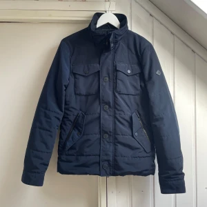 J. Lindeberg Jacka - Extremt snygg jacka från J.Lindeberg i en djupblå nyans med stilrena och funktionella detaljer. Storleken är S och är true to size. Jackan är lite tjockare och passar perfekt till hösten och vintern. Säljes för en billig peng till seriösa köpare.