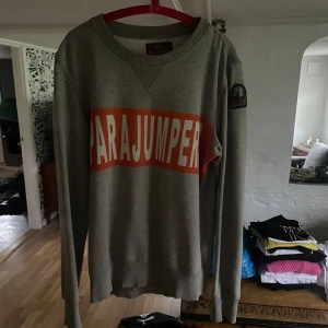 Grå tröja från Parajumpers - Säljer en snygg grå tröja från Parajumpers med ett stort orange tryck på framsidan. Tröjan är långärmad och perfekt för både höst och vår. Den har en normal passform och är superbekväm att ha på sig. Perfekt för en avslappnad stil!