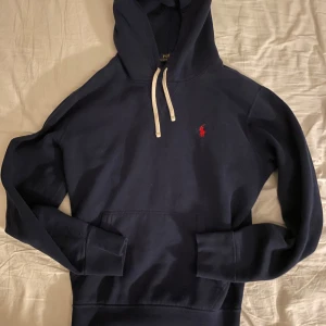 Mörkblå hoodie från Polo Ralph Lauren - Säljer en stilren mörkblå hoodie från Polo Ralph Lauren med det klassiska röda logotypbroderiet på bröstet. Tröjan har en justerbar huva med vita snören och en praktisk magficka. Perfekt för en avslappnad look. Mycket bra skick, inga defekter!