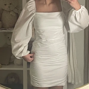 Vit klänning med puffärmar - Säljer en stilren vit klänning med puffiga långa ärmar och en fyrkantig urringning. Klänningen har en tight passform och är kort i modellen. Perfekt för en elegant look. Helt oanvänd 