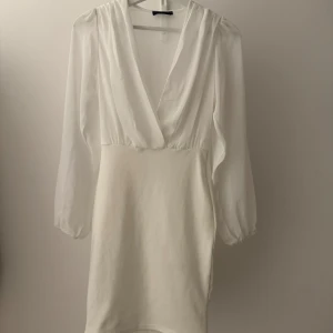 Vit klänning med v-ringning - Elegant vit klänning med v-ringad design och långa ärmar. Perfekt för en stilren look och STUDENTEN! Klänningen har en åtsittande passform i midjan och ett mjukt fall. Sitter super fint och formar kroppen helt magiskt! 