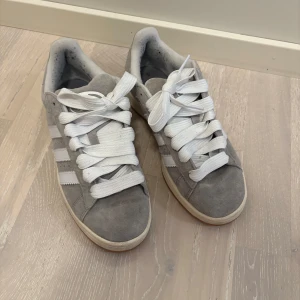 Grå Adidas Campus sneakers - Säljer ett par grå Adidas Campus sneakers i mocka med vita detaljer och snörning. Skorna har den klassiska tre-randiga designen på sidorna och en bekväm sula. Perfekta för en stilren och avslappnad look.