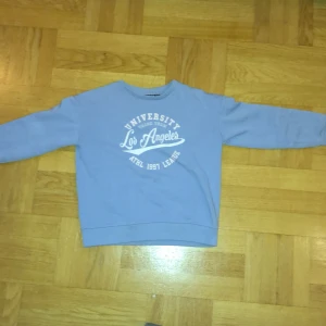 Blå sweatshirt från Reaction - Säljer en blå sweatshirt från Reaction med tryck 'University Los Angeles ATHL 1997 League' i vitt på framsidan. Tröjan har långa ärmar och en klassisk rund halsringning. Perfekt för en avslappnad stil!