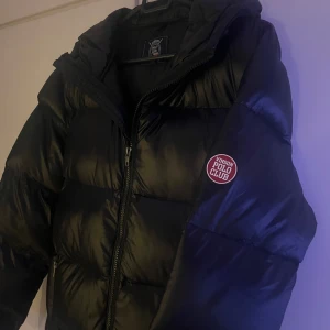 Svart pufferjacka från Vinson Polo Club - Snygg svart pufferjacka från Vinson Polo Club med dragkedja och huva. Jackan har ett stilrent märke på ärmen och är perfekt för kyligare dagar. Den är långärmad och har en glansig finish som ger en modern look. (Använd 1-2 gånger under vintern tills jag växte ur den)