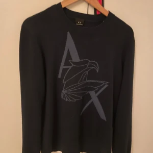 Svart tröja från Armani Exchange - Snygg svart tröja från Armani Exchange med ett stort tryck av en örn och bokstäverna 'A X' på framsidan. Tröjan har långa ärmar och en rund halsringning. Perfekt för en stilren look. Stl L men passar m