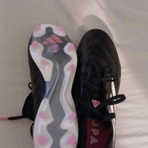 Svarta fotbollsskor från Adidas - Snygga svarta fotbollsskor från Adidas med vita och rosa detaljer. Skorna har snörning och en mönstrad sula med rosa dobbar för bra grepp på planen. Perfekta för fotbollsspelare som vill ha stil och funktion. Helt nya 