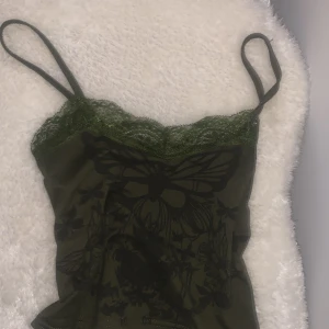 Grön linne med spetsdetaljer - Snygg grön linne med smala axelband och spetsdetaljer vid halsringningen. Linnet har ett mörkt fjärilsmönster.