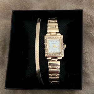 Guldfärgad klocka från Cartino + halsband  - Elegant guldfärgad klocka från Cartino med fyrkantig urtavla och glittriga detaljer runt kanterna. Klockan har en länkhalsband i metall och kommer i en snygg presentförpackning. Perfekt för att lägga till lite glamour till din stil.