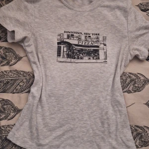Figursydd topp💓 - Superfin grå t-shirt/topp från Divided med ett fint tryck av en pizzeria i Downtown, New York. Perfekt för en avslappnad stil med en touch av storstadskänsla. Kortärmad och i mjukt material för extra komfort. Aldrig kommit till användning, pris kan alltid diskuteras!🫶🏼 köpt för runt 299 kr