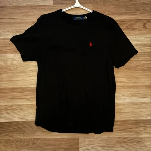 Svart t-shirt från Ralph Lauren - Snygg svart t-shirt från Ralph Lauren med det klassiska röda broderade logotypen på bröstet. Perfekt för en stilren och avslappnad look. Kortärmad och tillverkad i mjukt material för extra komfort. Stl Xl men passar även M