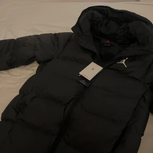 Svart dunjacka från Jordan - Säljer en snygg svart dunjacka från Jordan med huva och dragkedja. Perfekt för kyliga dagar. Jackan har en stilren design med en liten logga på bröstet. Helt ny i prislapp. Nyspris 2210kr
