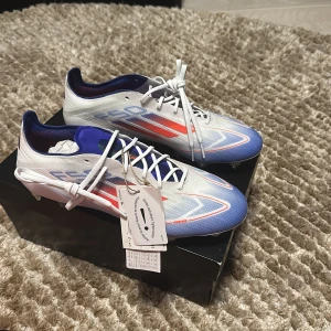 Vita och blå fotbollsskor från Adidas - Helt oöppnade, är från paketet. Fick de i fel storlek och kunde inte lämna tillbaka. Helt orörda med elite modell. Snygga fotbollsskor från Adidas i vitt med blå och röda detaljer. Skorna har snörning och en modern design som passar perfekt för fotbollsplanen. De har en strömlinjeformad form för optimal prestanda.