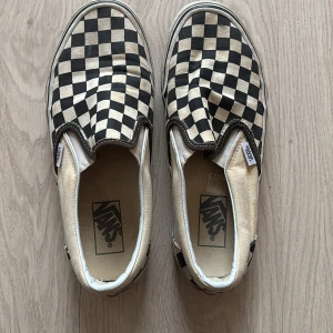 Svartvita slip-on från Vans - Säljer ett par klassiska slip-on sneakers från Vans med det svart/vit rutiga mönstret. Skorna har en bekväm passform och är i storlek 40l. De har en brun sula och är lätta att ta på och av. Säljer då de inte kommer till användning längre🙃🤗🫶