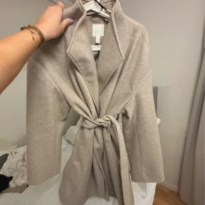 Beige kappa från H&M - Säljer en stilren beige kappa från H&M i storlek S. Kappan har en knytning i midjan och långa ärmar. Perfekt för höst och vår. Knappt använd.