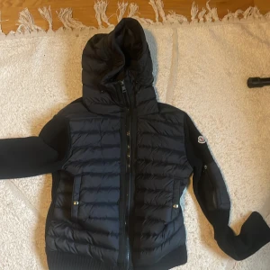 Svart cardigan från Moncler - Säljer en snygg svart pufferjacka från Moncler med huva och dragkedja. Jackan har ribbade detaljer och ett broderat Moncler-märke på ärmen. Perfekt för kyliga dagar!