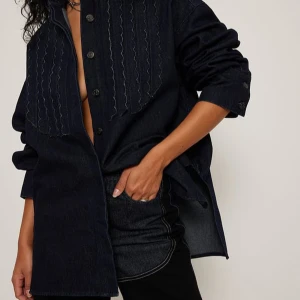 Mörkblå jeansskjorta - Snygg mörkblå jeansskjorta med knappar och dekorativa sömmar på framsidan. Skjortan har en loose passform och långa ärmar, perfekt för en avslappnad stil.