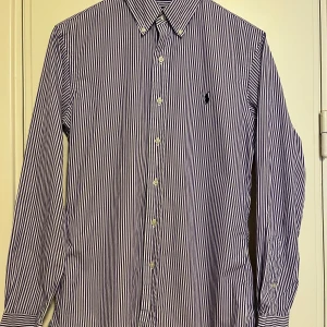 Randig skjorta från Ralph Lauren - Snygg randig skjorta från Ralph Lauren i lila och vit. Skjortan har en klassisk krage och knappar framtill. Den är i slim fit och tillverkad av bomull med stretch för extra komfort. Perfekt för en stilren look. Sparsamt använd i mycket gott skick. Se gärna mina andra annonser, så kan allt skickas i samma sändning. 