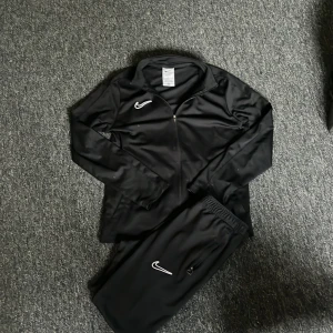 Svart träningsjacka från Nike - Säljer en svart träningsjacka från Nike med Dri-FIT-teknologi. Jackan har en dragkedja framtill och är långärmad. Perfekt för träning och sportiga aktiviteter. Den har en broderad Nike-logga på bröstet och är tillverkad i ett syntetmaterial för optimal komfort.