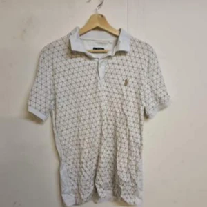 Vit pikétröja med geometriskt mönster - Säljer en vit pikétröja från Polo Ralph Lauren med ett brunt geometriskt mönster. Tröjan har korta ärmar och en klassisk krage. Perfekt för en stilren och avslappnad look.