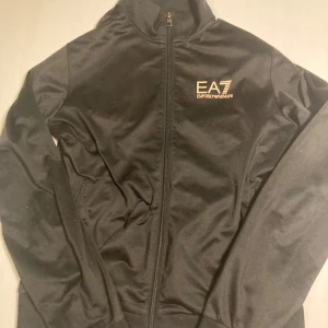 Svart tröja från Emporio Armani - Snygg svart tröja från Emporio Armani med dragkedja framtill och EA7-logga på bröstet. Tröjan har en stilren design med långa ärmar och en hög krage. Perfekt för en sportig look.