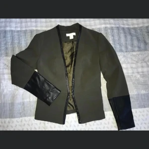 Olivgrön kavaj med svarta detaljer - Snygg olivgrön kavaj med svarta skinnliknande detaljer på ärmarna och längs kanterna. Perfekt för en stilren look. Kavajen har en öppen design utan knappar och är helfodrad för extra komfort.