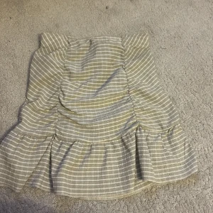 Randig kjol från Gina Tricot - Säljer en söt randig kjol från Gina Tricot i beige och vit. Kjolen har en volangdetalj längst ner och en tight passform som framhäver figuren. Perfekt för en stilren look.