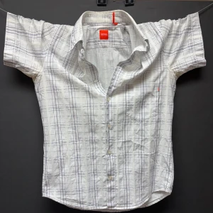 Vit rutig skjorta från Boss - Säljer denna snygga vita kortärmade skjorta från Hugo Boss. Najs mönster och fin detalj med det orangea. Perfekt för vår och sommar! Använts en del men är i väldigt bra skicka ändå!✅