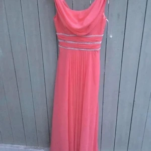 Korallfärgad långklänning med glittriga detaljer - Elegant korallfärgad långklänning med glittriga band i midjan. Klänningen har en draperad halslinje och är ärmlös, vilket ger en stilren look. Perfekt för speciella tillfällen.finns i norsborg botkyrka 