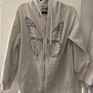 Grå hoodie med fjärilstryck - Säljer en grå hoodie med dragkedja och ett stort fjärilstryck på framsidan. Tröjan har en mysig huva med snören och praktiska fickor framtill. Perfekt för en avslappnad stil.