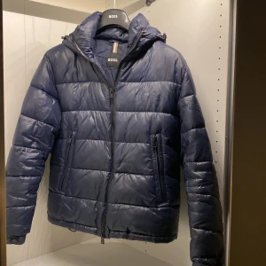 Mörkblå pufferjacka från Boss - Säljer en mörkblå pufferjacka från Boss med dragkedja och huva. Jackan har en quiltad design och är perfekt för kyligare väder. Den har en normal passform och är långärmad. Ord pris på jackan är 4500kr. Priset är förhandlingsbart vid snabb affär😃