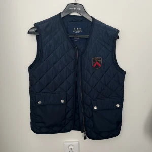 Morrisväst - Snygg mörkblå quiltad väst från Morris med dragkedja och två fickor med knappar framtill. Västen har en broderad logga på bröstet och är perfekt för lager-på-lager-stil. Västen är ärvd, men inte använd så jättemycket och har därför väldigt bra skick! Lite för liten på mig (185cm & 76kg). Bara att höra av sig angående frågor eller mer bilder🙌🏼