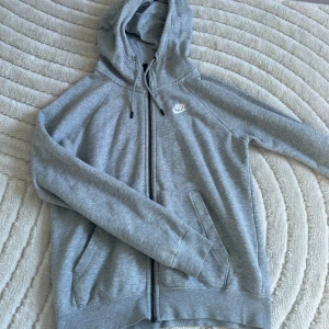 Grå hoodie från Nike - Säljer en grå hoodie från Nike med dragkedja och huva. Tröjan har långa ärmar och en liten vit Nike-logga på bröstet. Storlek XS men skulle säga S